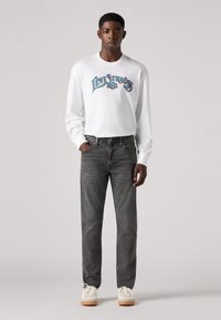 Cămașă albă cu mânecă lungă și logo colorat "Levi's", asortată cu blugi gri închis și teniși albi cu talpă maro deschis.