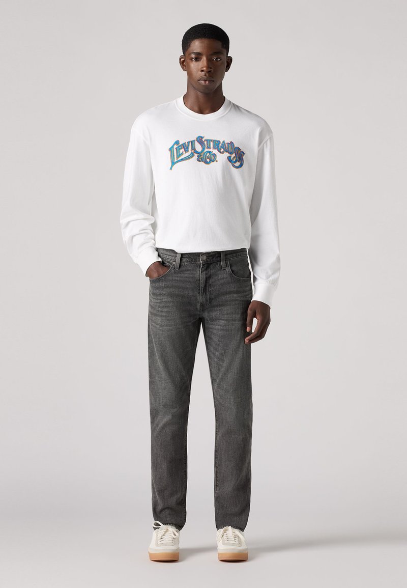 Chemise blanche à manches longues avec un logo "Levi's" coloré, associée à un jeans gris foncé et des baskets blanches avec une semelle marron clair.