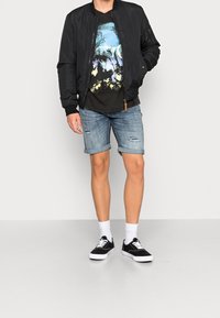 Mladý muž v černé bomber bundě, tričku s grafickým potiskem vlka, ohrnutých roztrhaných denimových šortkách, bílých ponožkách a černých teniskách na šedé podlaze.