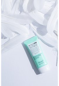 Mörkgrön tub av Benton UV Defense Sun Cream. Har vit text, ett flip-top lock och är omgiven av vita fjädrar på en vit bakgrund.