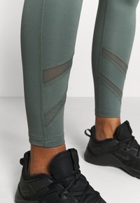 Legginsy sportowe w ciemnozielonym kolorze z siateczkowymi akcentami w geometryczny wzór. W połączeniu z czarnymi butami sportowymi. Gładka tekstura.