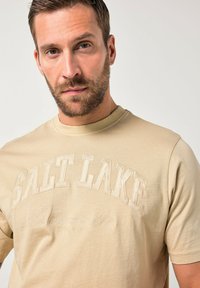 T-shirt beige in cotone con grafica testurizzata "SALT LAKE" in ricamo chiaro e scollo tondo con maniche corte.