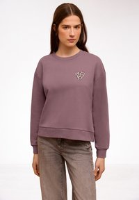 Femme portant un sweat-shirt mauve avec un cœur à imprimé léopard sur la poitrine et un jean gris délavé, debout devant un fond blanc.