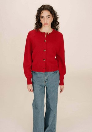 Cardigan en tricot rouge avec un col rond, des boutons en bois et un ourlet en côtes. Associé à un jean à jambes larges bleu clair.