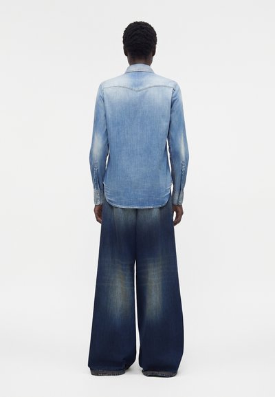 Άτομο που φορά ένα ανοιχτό μπλε τζιν πουκάμισο και φαρδιά σκουρόμπλε ξεθωριασμένα τζιν, στέκεται με την πλάτη προς την κάμερα σε απλό φόντο.