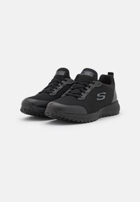 Schwarze Sportschuhe mit einer strukturierten Oberseite, flachen Schnürsenkeln und einer dicken Gummisohle. Mit Logo-Akzenten und einem eleganten Design.