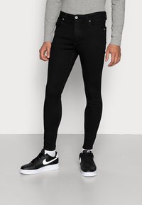 Jeans ajustados de mezclilla negra con una textura suave, diseño de cinco bolsillos, cierre de botón y costuras en contraste. Llevados con zapatillas negras.