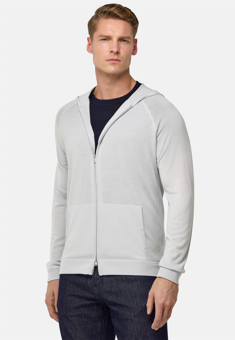 Sudadera con cremallera de color gris claro, hecha de tela suave, que dispone de un bolsillo frontal, puños acanalados y capucha con cordón. Llevada sobre una camiseta oscura.