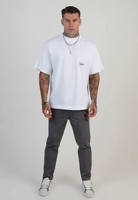 Witte katoenen T-shirt met een subtiel logo, gecombineerd met grijze cargobroek met zakken en zwart beslag. Afgewerkt met witte sneakers.