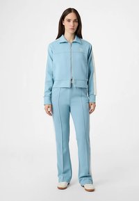 Lyseblå træningsdragt i glat stof. Jakke med lynlås og krave, med hvide sidelister. Tætsiddende bukser med sømdetaljer.