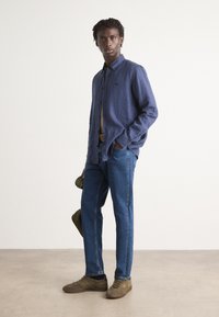Marine linbluse, lyseblå jeans og brune sneakers. Modellen står med hendene i lommene og holder en grønn hatt. Enkel bakgrunn.