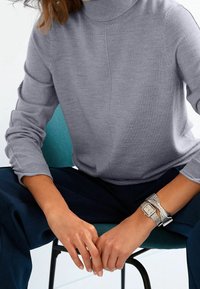 Pull en col roulé gris avec des manches longues, texture tricotée et poignets côtelés. Montre métallique avec un bracelet tressé au poignet.