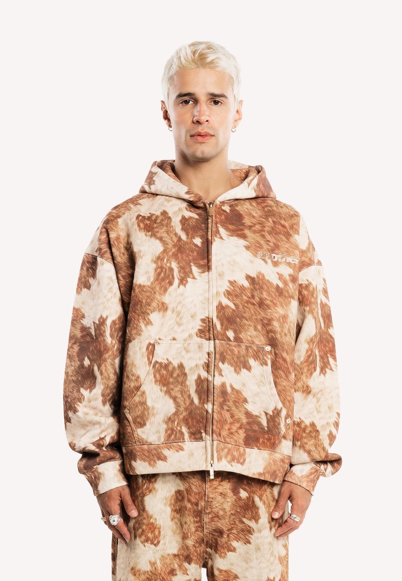 Jeune personne aux cheveux blonds courts portant un sweat à capuche zippé imprimé vache marron et blanc avec un pantalon assorti, debout devant un fond uni.