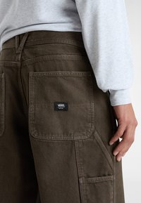 Bruna cargobyxor med stora fickor, rak passform och logotygmärke. Bärs med en ljusgrå sweatshirt med lång ärm.