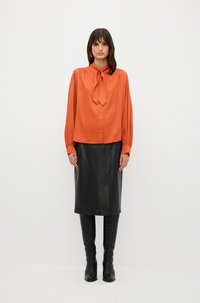Orange-satin Bluse mit Halstuch, Knopfverschluss und langen Ärmeln, kombiniert mit einem schwarzen Leder-Midikleid und kniehohen schwarzen Stiefeln.