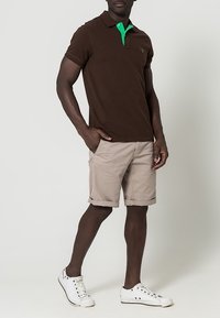 Polo marron avec un col vert, assorti à un short beige. Le mannequin porte des baskets blanches. Coupe décontractée, manches courtes, sans motifs visibles.
