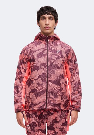 Homme portant une veste à capuche zippée à motifs roses et bordeaux et un short assorti, debout face à l'avant sur un fond uni.