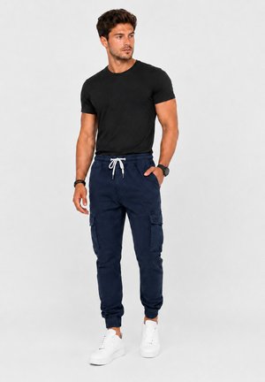 Mann trägt schwarzes kurzärmeliges Shirt, marineblaue Jogginghose mit Seitentaschen, weiße Sneakers und Armbanduhr, steht vor einfarbigem Hintergrund.