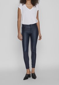 Pantalones de imitación cuero en color marino con un diseño ajustado, combinados con una camiseta blanca de cuello en V. La modelo lleva mules negros de tacón. Textura suave.