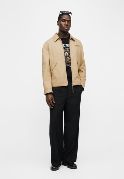 Άνδρας που στέκεται, φοράει μπεζ μπουφάν πάνω από μαύρο μπλουζάκι με τύπωμα αρκούδας, μαύρο παντελόνι φαρδιάς γραμμής, μαύρα παπούτσια και γυαλιά ηλίου στο κεφάλι.