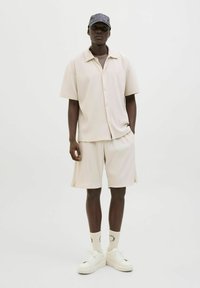 Beige csíkos, rövid ujjú ing és hozzáillő shorts, fehér sportcipővel és mintás zoknival párosítva. A modellen sötét mintás sapka van.