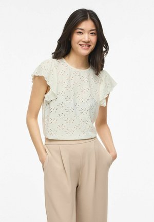 Młoda kobieta uśmiechająca się, ubrana w białą bluzkę z krótkim rękawem i ażurowym wzorem oraz beżowe spodnie o szerokich nogawkach, z rękami w kieszeniach.
