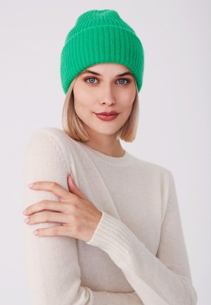 Beanie verde a coste con vestibilità aderente. Abbinato a un maglione a maniche lunghe di un beige chiaro, con un motivo testurizzato sui polsini.