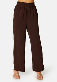 Bruine, high-waisted, wijdvallende broek met een gathered elastische tailleband; lichte, gestructureerde stof; gecombineerd met gevlochten slidersandalen.