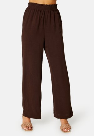 Pantaloni marroni a vita alta, a gamba larga, con una cintura elastica arricciata; tessuto leggero e testurizzato; abbinati a sandali a ciabatta intrecciati.