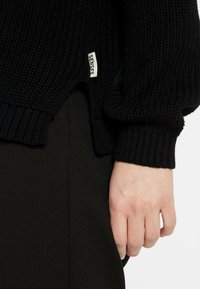 Schwarzer Strickpullover mit einem strukturierten Muster, ausgestattet mit gerippten Bündchen und einem seitlichen Etikett mit der Aufschrift "SENSE". Kombiniert mit dunklen Stoffhosen.