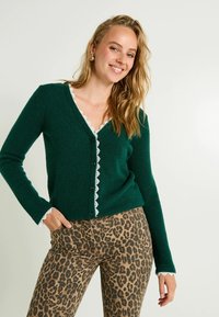 Cardigan vert avec un décolleté en V profond, bordure en dentelle blanche et fermeture à boutons. Porté avec un pantalon léopard marron. Texture douce et design ajusté.