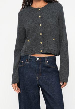 Kvinde iført mørkegrå knap-ophængt cardigan med forlommer og høj talje bredbenede mørkeblå jeans, stående mod en hvid baggrund.