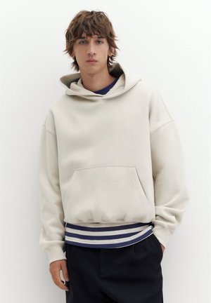 Un jeune homme porte un sweat à capuche oversize couleur crème, à l’ourlet rayé, et un pantalon noir sur un fond blanc uni.