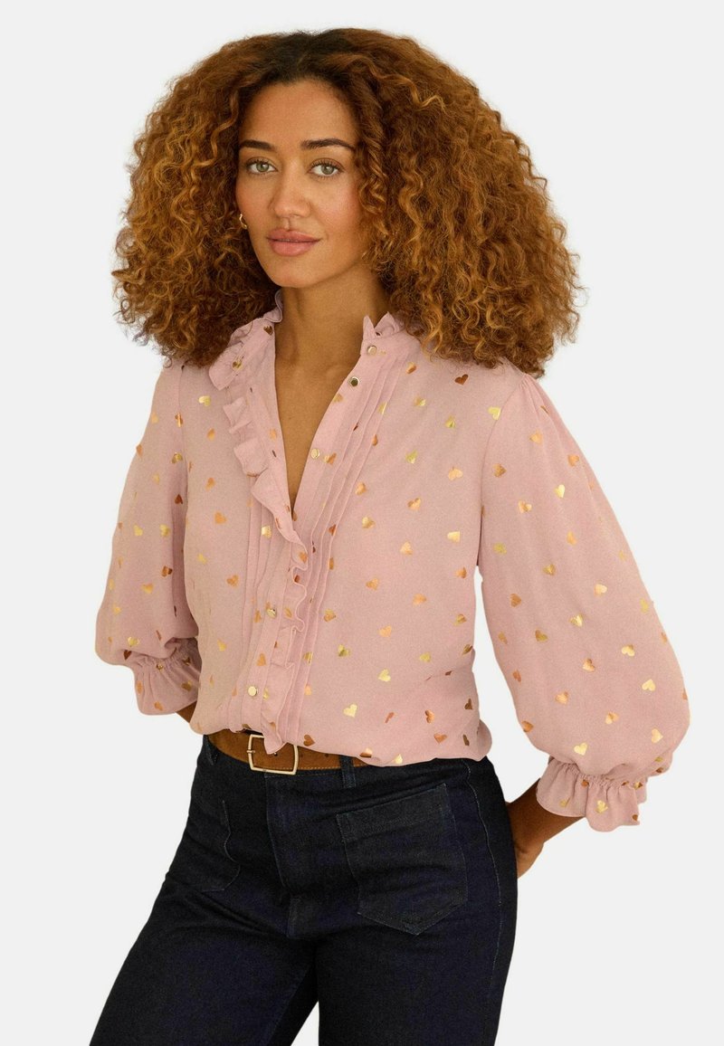 Naine mahuliste lokkis juustega, kandes roosat pluusi kuldsete südamekujundustega ja tumedaid kõrge vöökohaga teksaseid, poseerib käed selja taga.