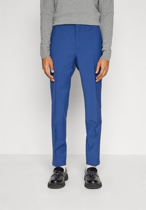 Pantaloni sartoriali blu con una texture liscia, caratterizzati da una parte frontale piana e una piega. Abbinati a un maglione grigio e scarpe nere.