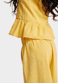 Žlutý lněný dvoudílný outfit se skládá z peplum topu a kalhot s elastickým pasem, zdůrazňuje texturovanou tkaninu a ležérní siluetu.