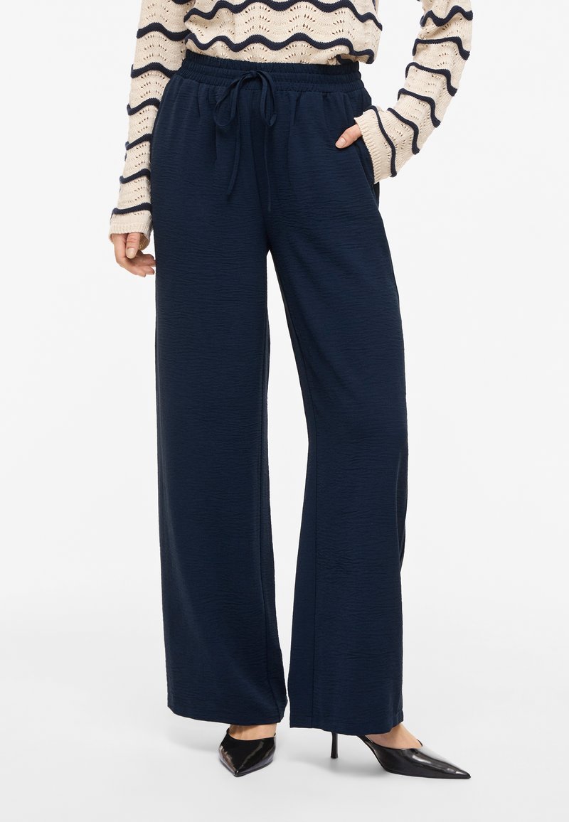 Κυανές παντελόνες με πλατιά γραμμή, ελαστική μέση και κορδόνι, φορεμένες με κρεμ πουλόβερ με κυανές κυματιστές ρίγες και μαύρες μυτερές γόβες.