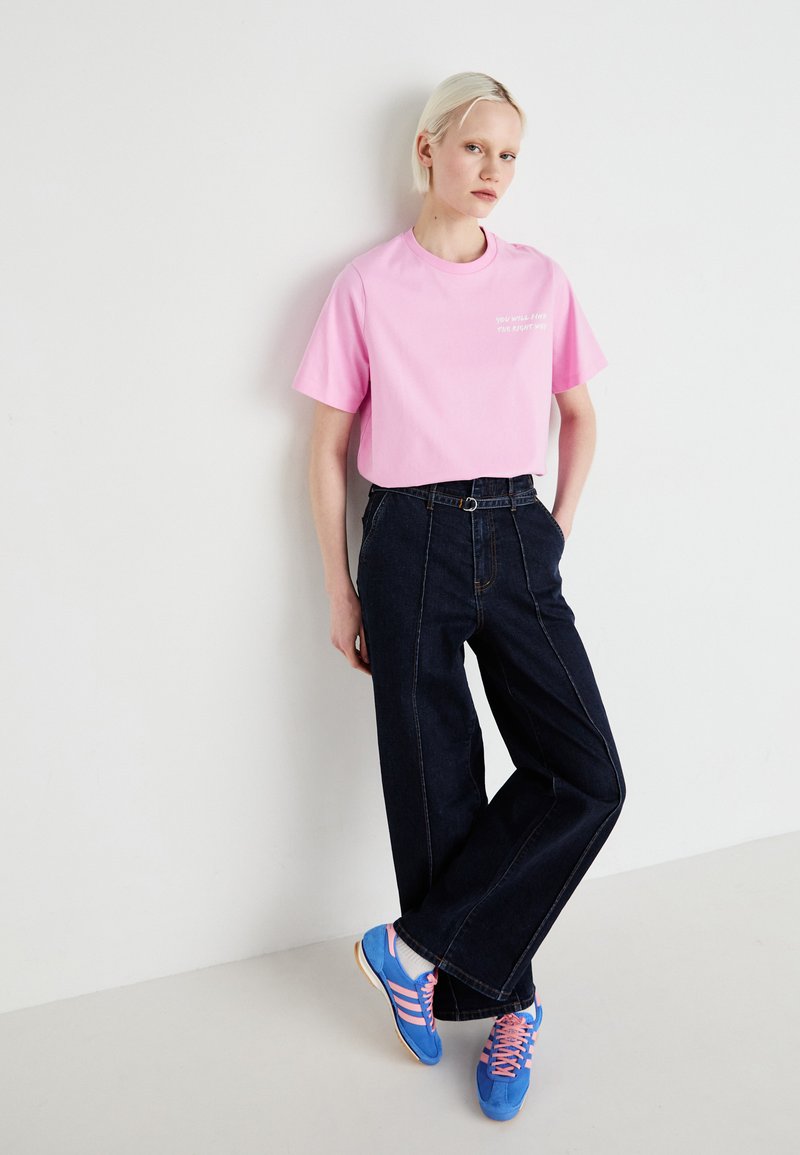 Roze katoenen T-shirt met korte mouwen, gecombineerd met hooggerende donkere spijkerbroek. Blauwe sneakers met roze accenten op een lichte achtergrond.