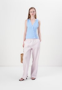 Cardigan sin mangas azul con botones, combinado con pantalones de pierna ancha a rayas color rosa claro. La modelo sostiene un bolso de mimbre. Diseño simple, material suave.