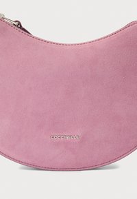 Roze suède tas in halvemaanvorm met een gladde textuur, voorzien van een zilveren logo aan de onderkant en een rits aan de bovenkant.