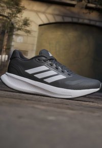 Chaussure de sport gris foncé avec une semelle blanche et trois bandes blanches sur le côté, reposant sur une surface en pierre à l'extérieur.