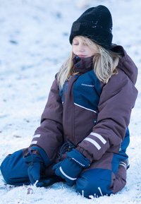 Barn som bär en brun och blå vinterjacka med vita ränder, marinblå handskar och en svart mössa, sitter på snötäckt mark.
