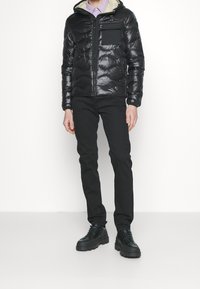 Veste matelassée noire avec capuche, dotée d'une fermeture éclair et d'une poche sur la poitrine. Associée à un pantalon slim noir et des bottes noires épaisses.