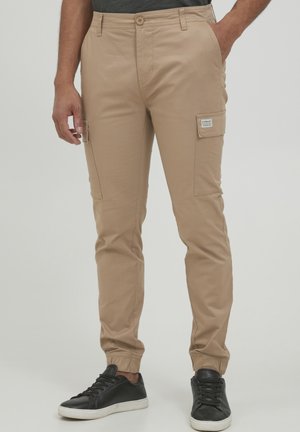 Pantalones cargo beige con corte ajustado, que cuentan con bolsillos laterales y puños elásticos. Fabricados con un tejido suave y duradero. Combinados con zapatillas de deporte negras.