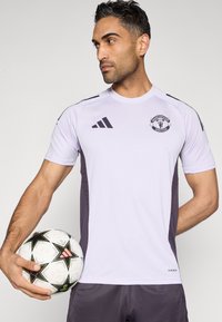 Purpurinės ir pilkos spalvos sportinis marškinėlis trumpomis rankovėmis, ant krūtinės - „Manchester United“ logotipas. Žaidėjas laiko raštuotą futbolo kamuolį.