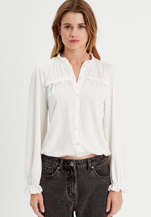Witte blouse met lange mouwen, voorzien van een textuur patroon, knoopsluiting aan de voorkant, rimpelaccenten aan de halslijn en borst, en gathered manchetten.