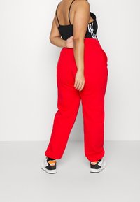 Person klädd i röda sweatpants, svart toppen med remmar och vita Adidas-sneakers som står med ryggen vänd mot kameran framför en enkel vit bakgrund.