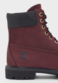 Bordeauxrotes Wildleder-Ankle-Boot mit einem schwarzen Lederkragen, goldenen Ösen, schwarzen Schnürsenkeln und einem schwarzen Baumlogo an der Seite. Texturiertes Gummisohle.