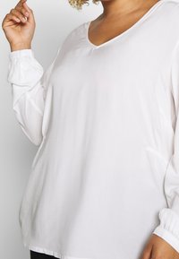 White long-sleeve top with a V-neck, loose fit, and gathered sleeves. Soft, smooth fabric with subtle sheen and no visible patterns.