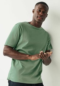Camiseta de algodón verde con escote redondo y mangas cortas, de textura suave, combinada con jeans oscuros, manos juntas al frente.